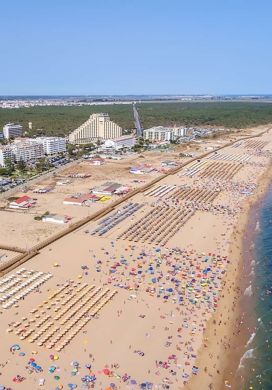 Praia de Vila Real de Santo António, com extenso areal, mar azul e edifícios ao fundo, incluindo hotéis e apartamentos