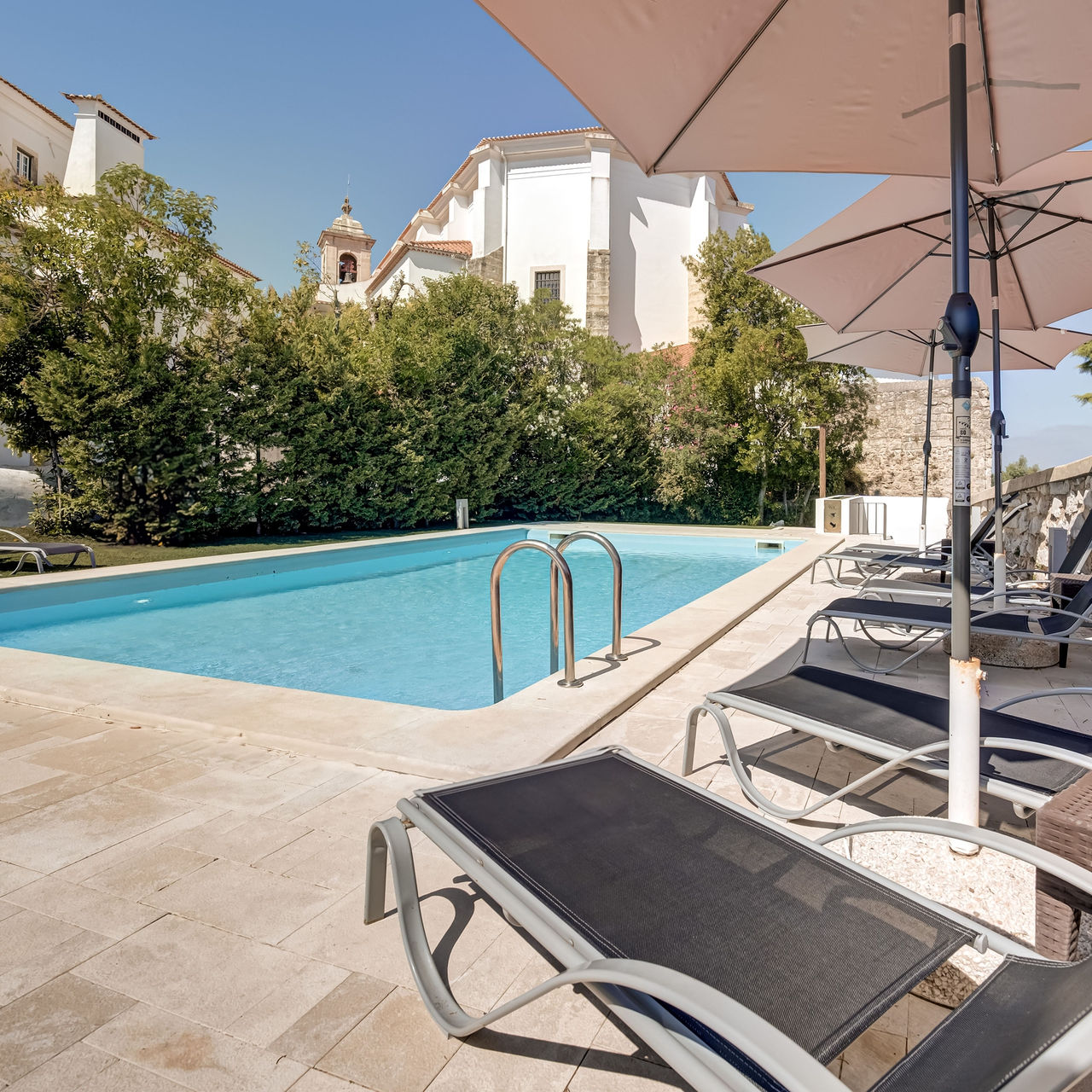 Piscina exterior da Pousada Ourém, um hotel de charme perto de Fátima, com espreguiçadeiras, chapéus de sol e várias árvores