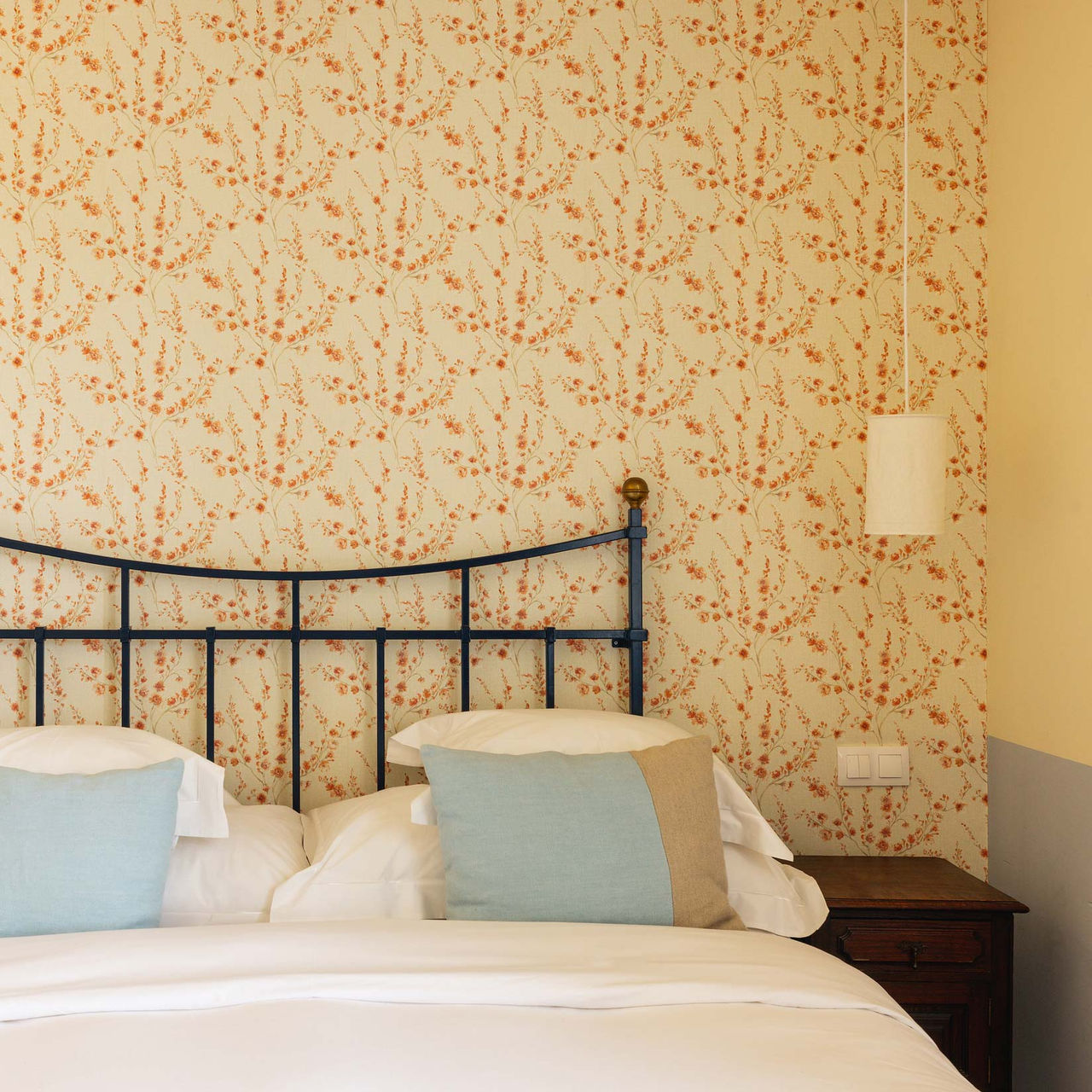 Cama com cabeceira de ferro, almofadas em tons claros e parede com papel de flores na Pousada Vila Óbidos