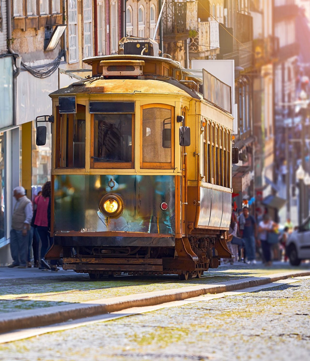 Vista de um elétrico amarelo antigo a subir uma rua na cidade do Porto, com várias pessoas atrás e edifícios antigos