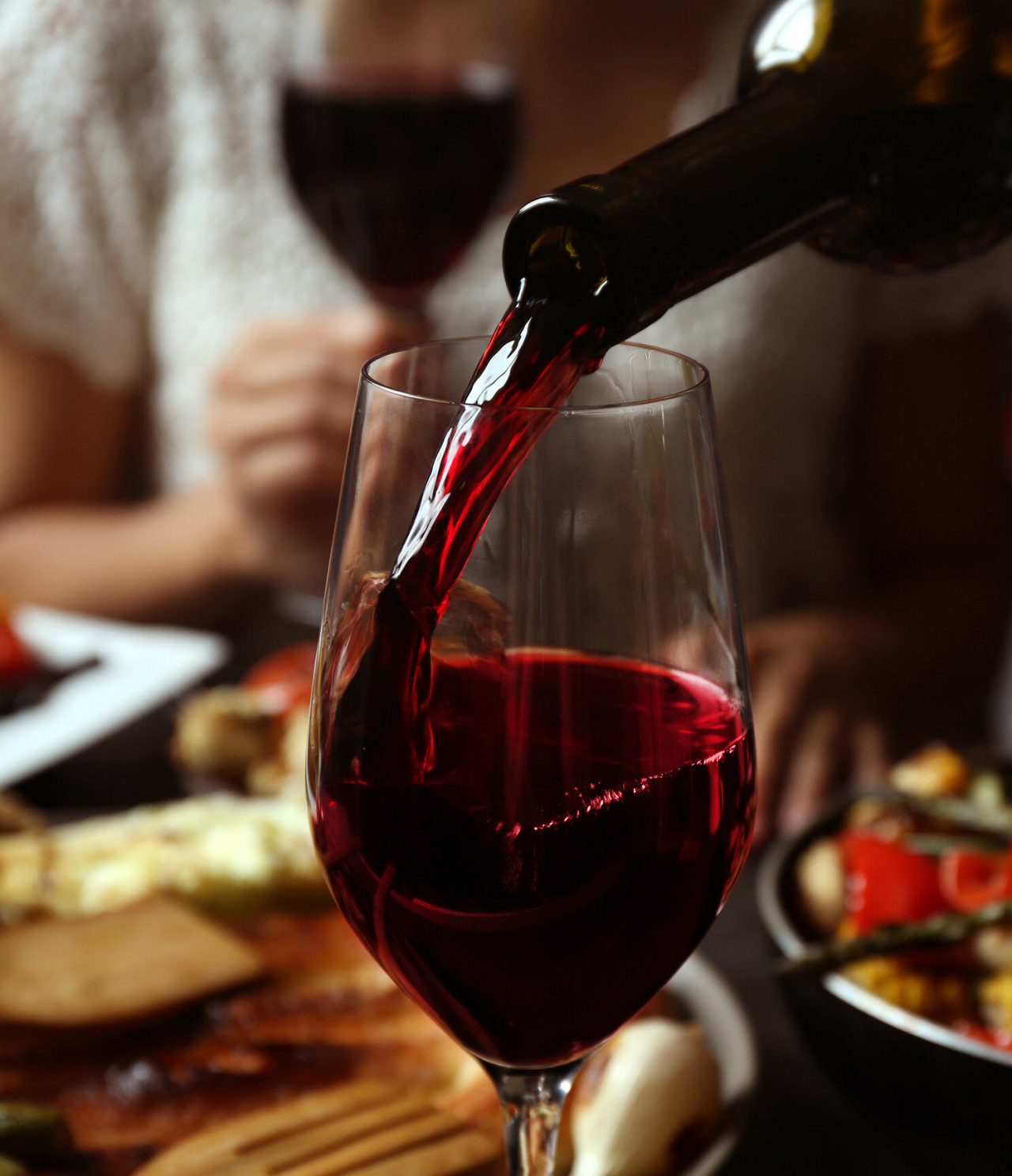 Vinho tinto do Porto a ser servido num copo alto, com uma pessoa atrás a beber vinho e pratos de comida