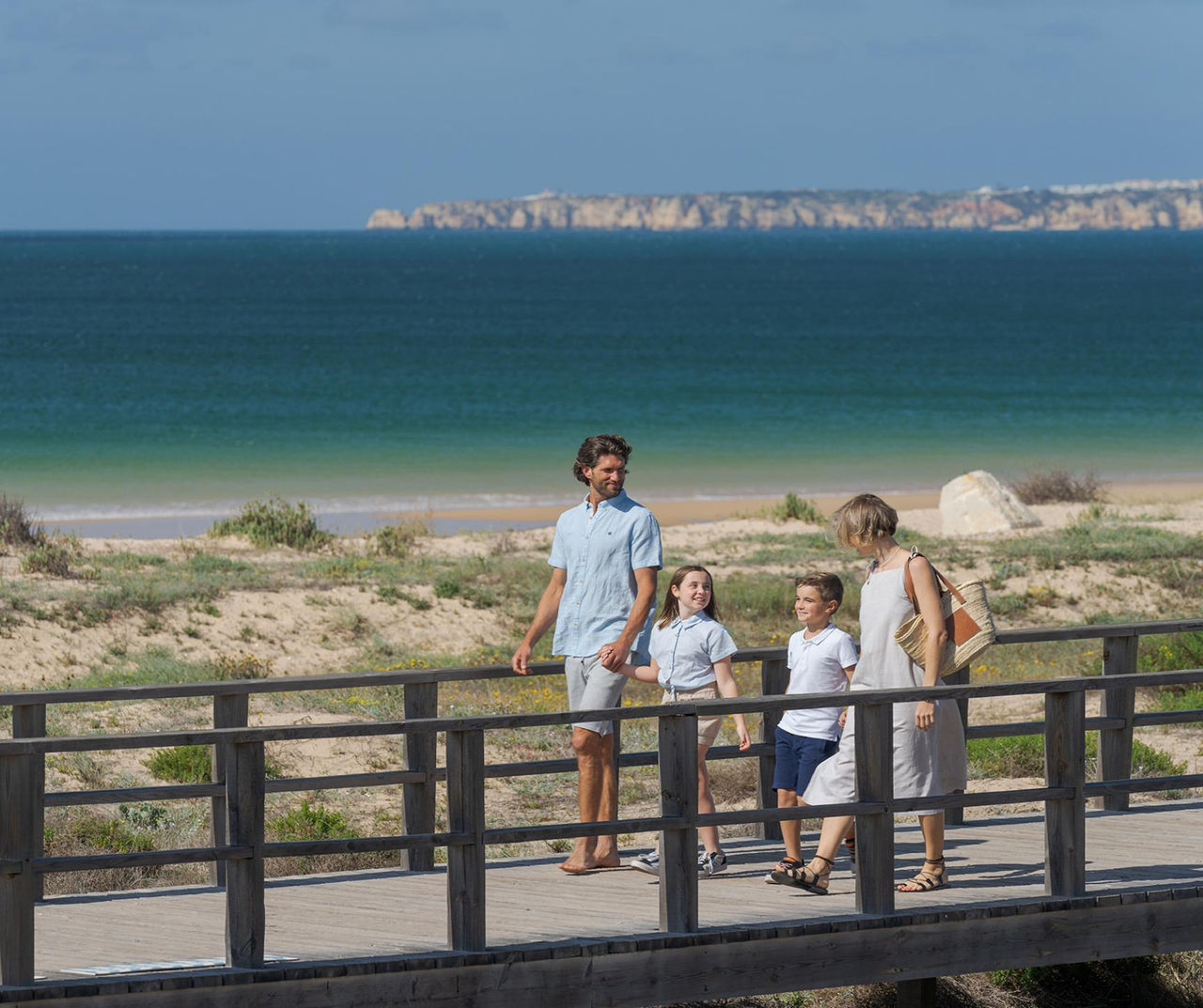 Hotel Algarve onde famílias passeiam num passadiço junto à praia com vista para o mar azul e falésias
