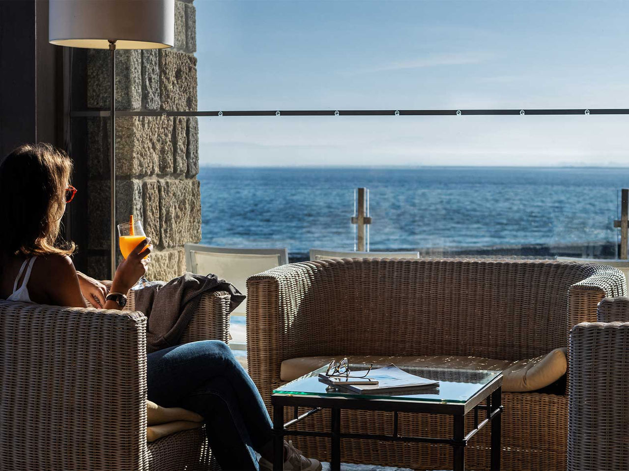 Mulher sentada em poltrona de vime com bebida, vista para o mar através de janela de vidro