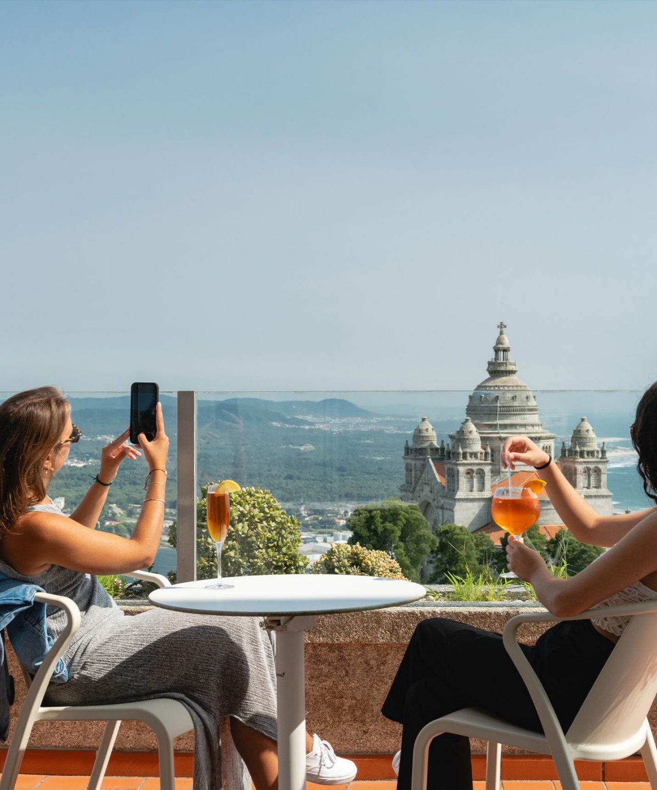 Duas pessoas sentadas num terraço com bebidas, apreciando vista para igreja e colinas ao fundo