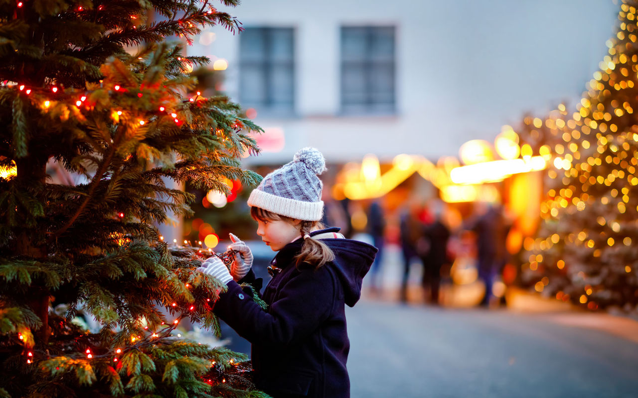 Menina com gorro de inverno a decorar uma árvore iluminada num mercado de Natal ao entardecer com luzes desfocadas