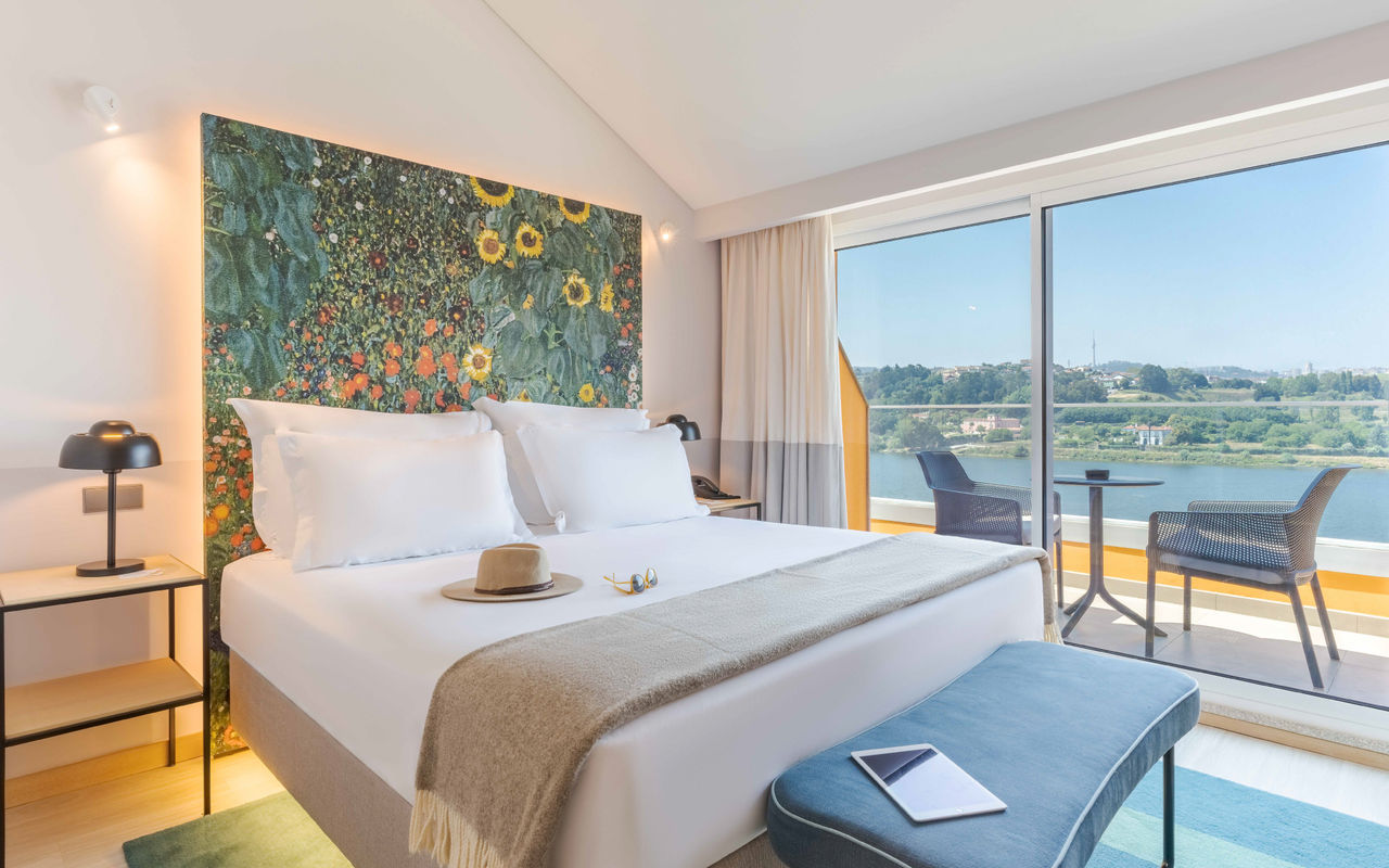 Quarto luminoso com cama de casal, varanda com vista rio, cadeiras exteriores e painel floral colorido