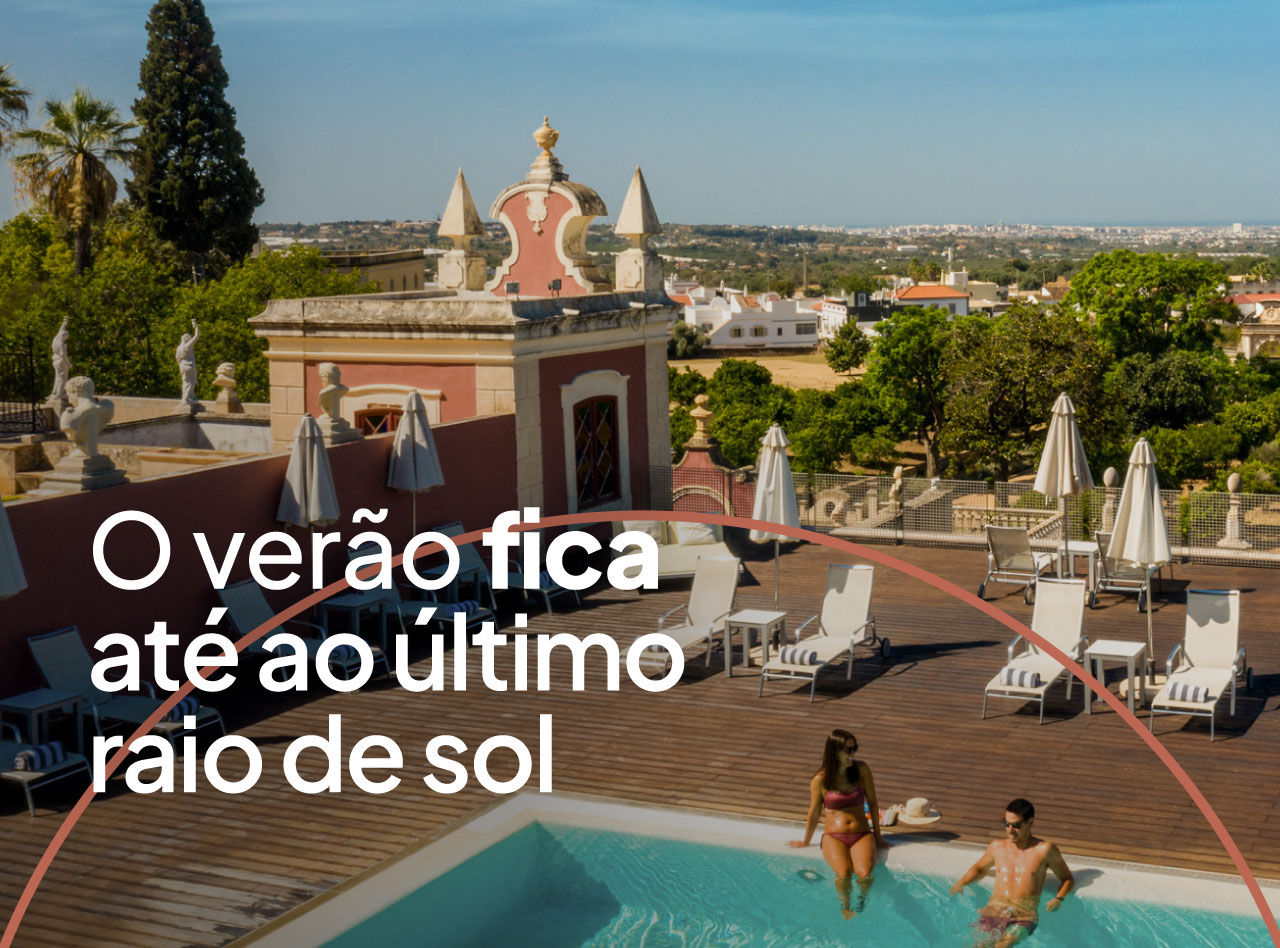Casal relaxa numa piscina sobre deck de madeira junto a edifício histórico e vista ampla sobre árvores