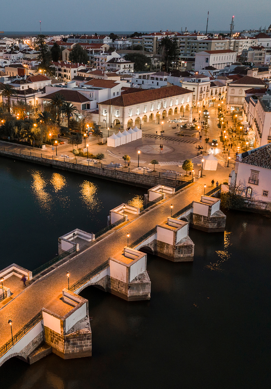 Typical view of Tavira with a Roman bridge over the Gilão River at sunset with lights illuminating the square