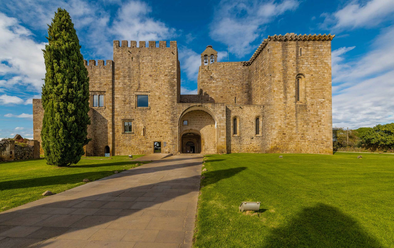 Historic stone castle with arched entrance, green lawns and tall cypress tree beneath a blue cloudy sky