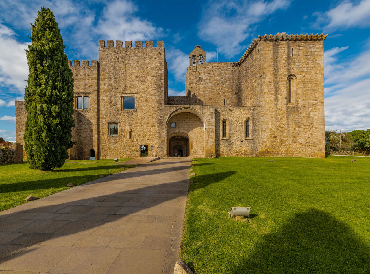 Historic stone fortress with arched entrance, green lawns and tall cypress tree beneath a blue cloudy sky